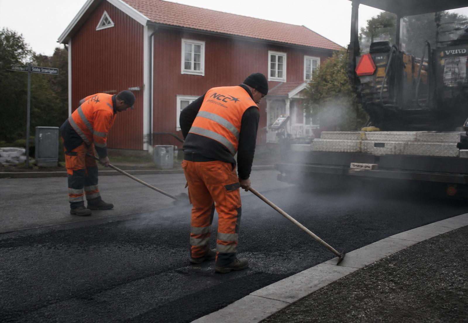 Två asfaltsarbetare i orange varselkläder räfsar het asfalt bakom utläggaren, framför en Falu-röd träfasad i Jönköping